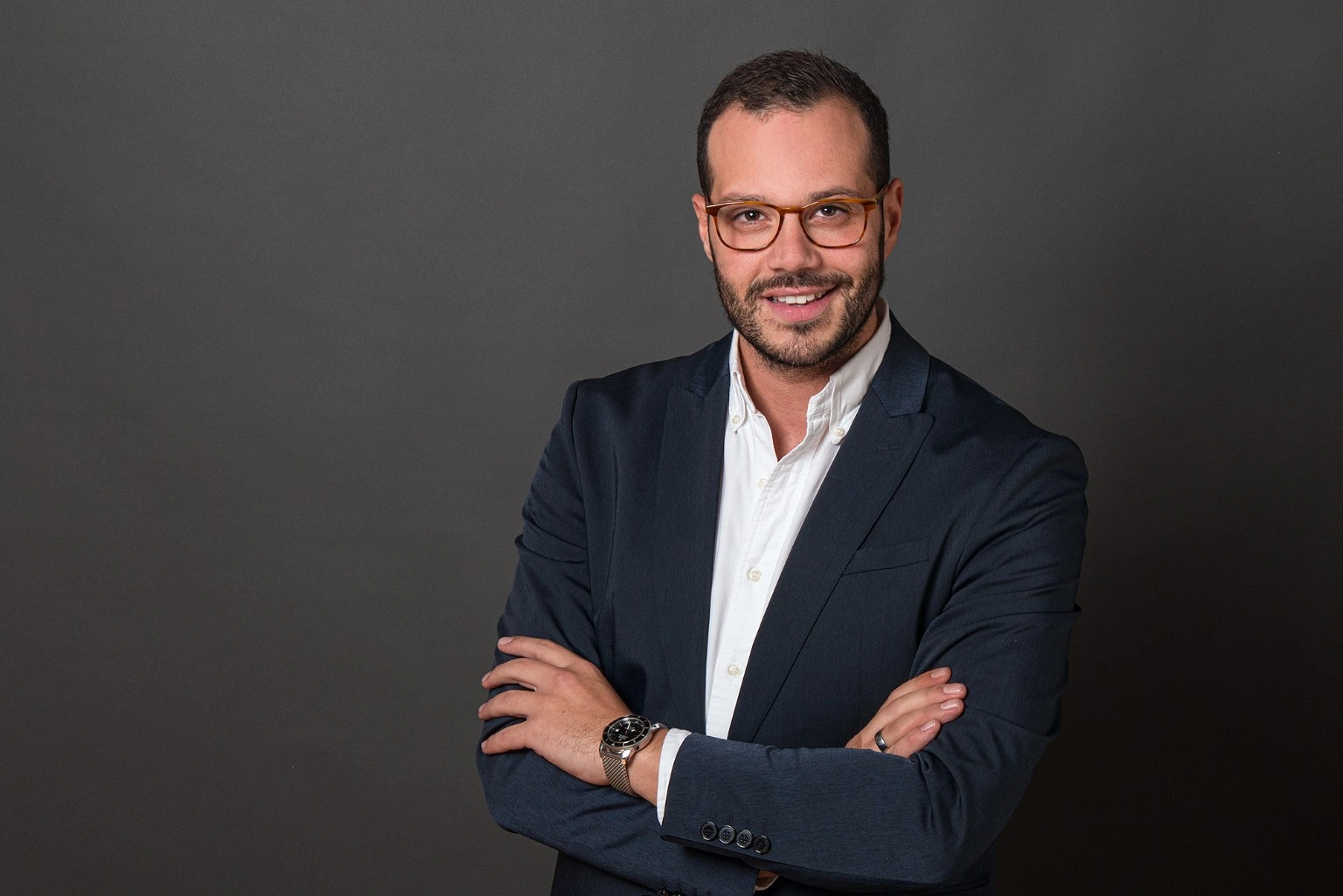 Bewerbungsfoto eines Mannes mit Brille und verschränkten Armen im dunklen Sakko